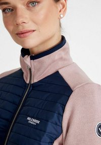 Giacca blu navy e rosa chiaro con design trapuntato, colletto alto e chiusura a zip. Presenta una toppa del logo sulla spalla e una texture liscia.