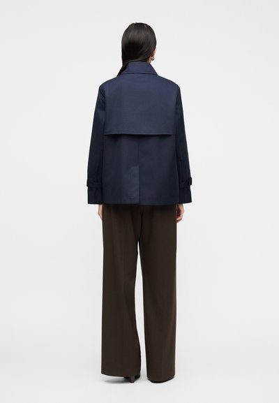 Γυναίκα που φοράει ένα ναυτικό cropped σακάκι με λεπτομέρεια πίσω στον ώμο και καφέ παντελόνι φαρδιάς γραμμής, στέκεται με την πλάτη προς τα έξω σε απλό φόντο.