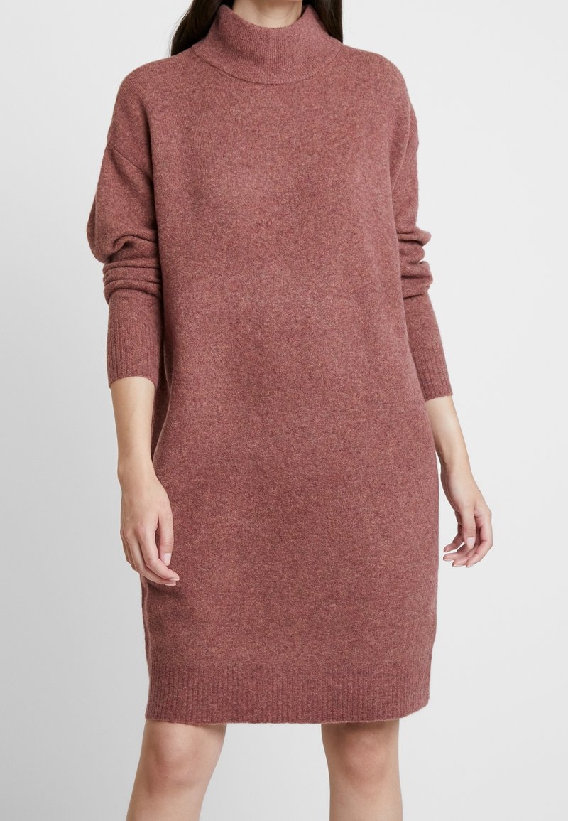 Femme portant une robe-pull ample couleur mauve à manches longues et longueur aux genoux avec un col montant sur un fond uni.