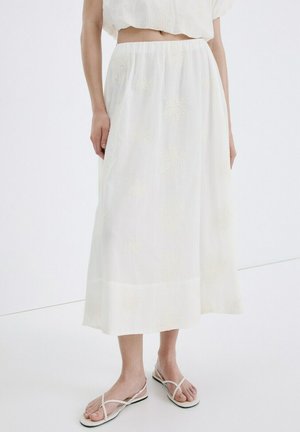 Falda midi blanca bordada combinada con un top a juego y sandalias planas blancas con tiras, en una persona de pie sobre un fondo liso.