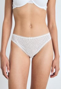 Hvitt blondeundertøy med geometrisk mønster, tynne scallopkanter og en liten sløyfedetalj foran. Mykt, delikat materiale.