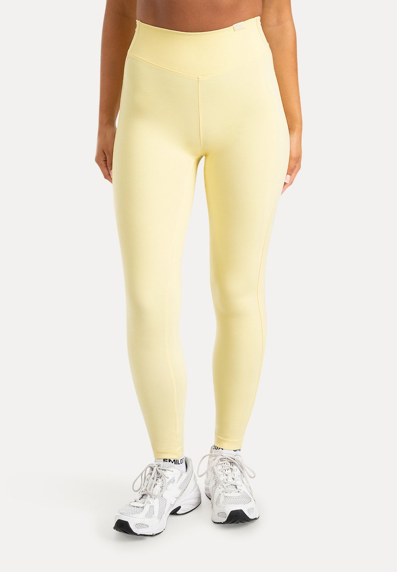 Femme portant des leggings jaune pâle à taille haute et des chaussures de sport blanches, debout devant un fond uni.