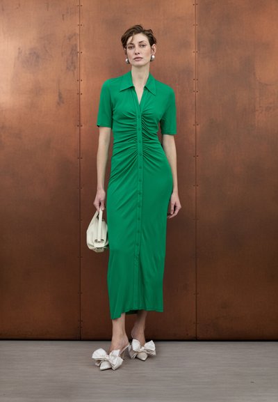 Vestido ajustado verde con mangas cortas y botones delanteros. La tela texturizada se complementa con zapatos de tacón blancos adornados con lazos y un bolso color crema.