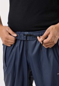 Laivastonsiniset urheilushortsit, jotka on valmistettu kiiltävästä, kevyestä materiaalista. Niissä on joustava vyötärönauha ja musta kiristysnauha säädön helpottamiseksi.