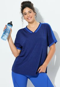 Uśmiechnięta kobieta w niebieskim stroju sportowym trzyma w jednej ręce butelkę z wodą z napisem „VAUDE”, pozując na białym tle.