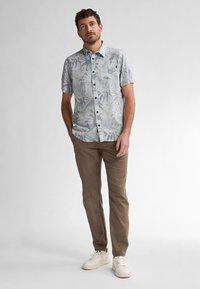 Camicia a maniche corte, di colore azzurro chiaro con motivo a foglie, con bottoni sul davanti e taschino sul petto, abbinata a pantaloni marroni chiari e sneakers bianche.