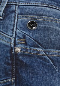 Granatowe dżinsy z miedziowanym przeszyciem, czarnym, okrągłym guzikiem z logo "MAVI" oraz metalowymi nitami na kieszeni.
