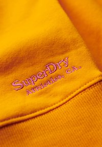 Un tejido amarillo brillante presenta el texto bordado "SuperDry Athletics, CA" en color rosa. La textura es suave con un detalle de costura ligeramente elevado.