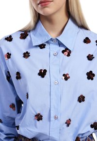 Lyseblå skjorte med knapper, der har mørke blomsterpailletter spredt over stoffet. Blød tekstur med klassisk krave og knaplukning.