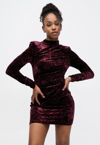 Robe courte en velours de couleur bordeaux profond avec des manches longues et un col montant. Présente des fronces le long du corps pour ajouter de la texture et de la forme.