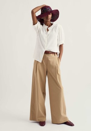 Femeie purtând o bluză albă cu nasturi, pantaloni bej cu picioare largi și curea maro, pălărie lată bordo și pantofi asortați, stând cu o mână ridicată.