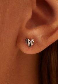 Boucle d'oreille en argent en forme de cœur, avec une texture ondulée, de petite taille et une finition polie, positionnée près de l'oreille.