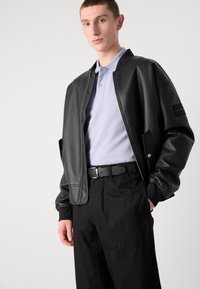 Black faux leather bomber jacket with a zip front, ribbed cuffs, and side pockets, worn over a light purple polo and black trousers.