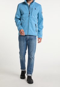 Uomo che indossa una giacca azzurra leggera con zip, jeans blu con risvolti e stivali neri, in piedi su uno sfondo bianco.