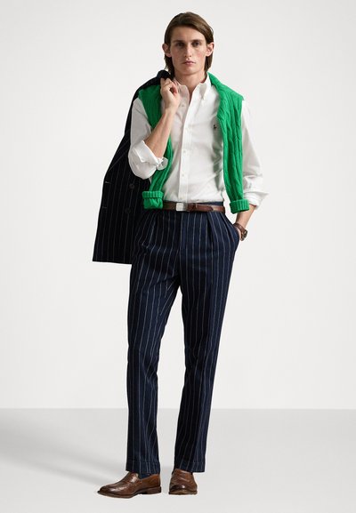 Gilet matelassé vert, chemise blanche à boutons, pantalon à rayures bleu marine, chaussures en cuir marron, et un blazer sombre drapé sur l'épaule.
