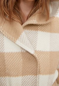 Béžový a bílý kostkovaný vlněný kabát s velkými klopami, který nosí osoba s červenými vlasy.