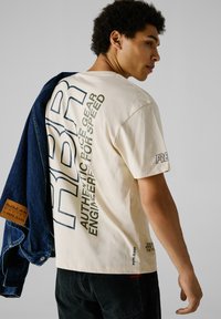 Camiseta de algodón beige con texto gráfico en la espalda y en la manga, combinada con una chaqueta de denim y jeans negros.