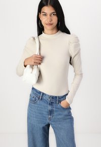 Bluza lungă de culoare bej deschis cu mâneci bufante, asociată cu blugi albaștri cu talie înaltă și o poșetă mică albă.