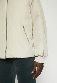 Veste beige à fermeture éclair avec poche zippée latérale portée sur un pantalon sombre, montrant une main à la peau foncée reposant le long du corps.