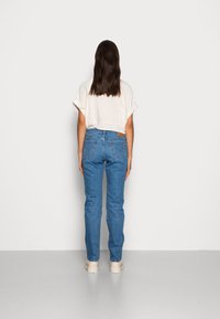 Jeans de ganga azul com unificação reta e design de cinco bolsos, combinados com uma blusa curta de cor clara. Costuras visíveis e etiqueta de couro na parte de trás.