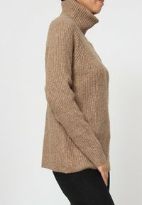 Pull à col roulé beige à côtes avec des manches longues et une coupe décontractée. Présente un matériau tricoté texturé et doux. Ourlet légèrement asymétrique.