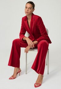 Rød fløjlssæt med en tætsiddende blazer og brede bukser, parret med lyserøde strophæle. Den glatte tekstur og den dybe farve skaber et dristigt look.