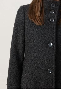 Mørkegrå uldfrakke med tekstureret overflade, høj krave og seks store knapper foran. Lange ærmer fuldender designet.