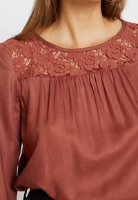 Blusa color ruggine con yoke in pizzo, parte frontale plissettata e maniche lunghe. Tessuto liscio con dettagli in pizzo floreale sul colletto.