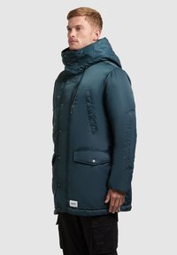 Tmavě modrý voděodolný parka s vysokým límcem, kapucí, dvěma bočními kapsami a jednou kapsou na hrudi. Obsahuje patentky a hladký povrch.