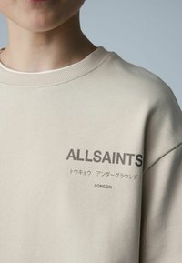 Sweatshirt beige avec col rond, col côtelé et logo "ALLSAINTS" en gris foncé accompagné d'un texte en japonais et de "LONDON".