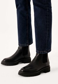 Black leather Chelsea boots with elastic side panels, chunky textured soles, and rounded toes, worn with dark denim jeans.