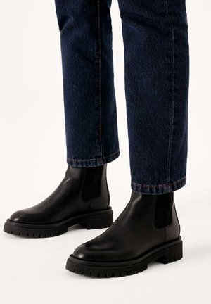 Black leather Chelsea boots with elastic side panels, chunky textured soles, and rounded toes, worn with dark denim jeans.