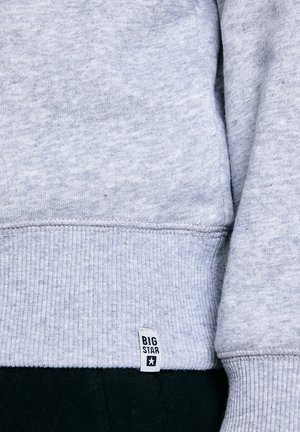 Světlé šedé manžety žebrované mikiny s bílou visačkou označenou "BIG STAR" nad černými kalhotami.