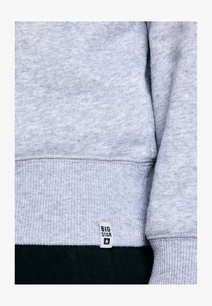 Helehall triibuline dressipluusi varrukas valge sildiga, millele on peale trükitud "BIG STAR", mustade pükste kohal.