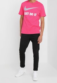 Rosa Nike t-shirt med grå "JUST DO IT" text, korta ärmar; kombinerad med svarta byxor och vita sneakers. Enkelt design, avslappnad passform.