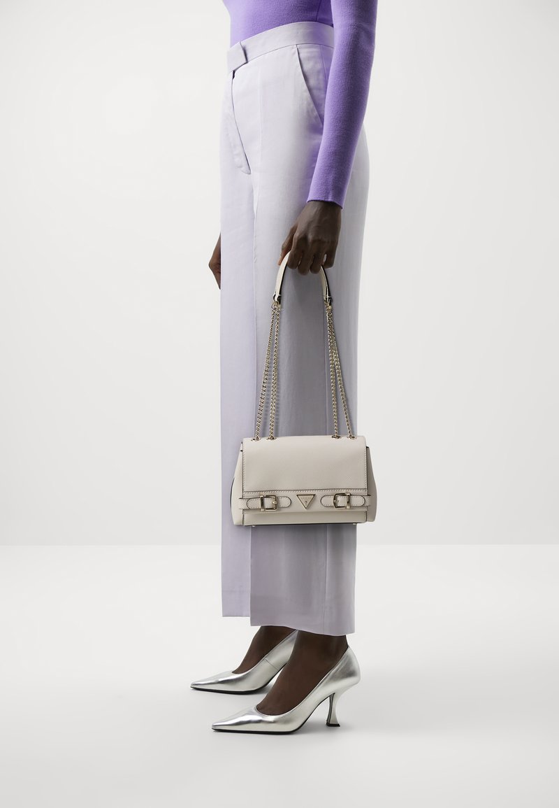 Borsa in pelle beige con tracolla a catena, logo triangolare e dettagli con fibbia. Il modello indossa pantaloni larghi lavanda e tacchi silver.