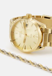 Goldfarbener Metallarmbanduhr mit rundem Zifferblatt, auffälligen Stundenmarkierungen und Datumsanzeige. Mit einer geflochtenen zweifarbigen Kette.