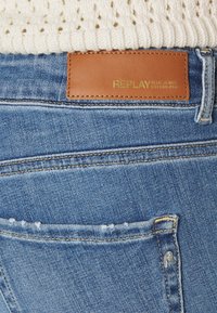 Blå denimjeans med en brun läderetikett märkt "REPLAY." Har en blekt tvätt, kontrasterande sömmar och minimal slitning.