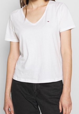 Kobieta ubrana w białą koszulkę z krótkim rękawem i dekoltem w serek z małym logo Tommy Hilfiger na piersi oraz czarne spodnie.