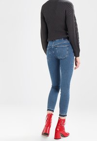 Kvinna iklädd en mörkgrå stickad tröja, blå skinny jeans och röda lackskinnankelboots med runda dragkedja, stående med ryggen mot kameran.