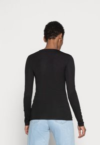 Persona con capelli ricci e corti che indossa un top nero a maniche lunghe aderente e jeans blu chiaro, di spalle contro uno sfondo neutro.