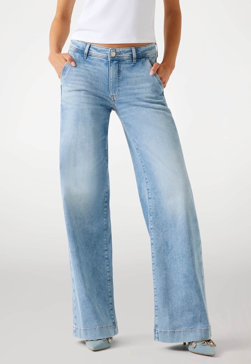 Personne portant un jean bleu clair à jambes amples, les mains dans les poches et des chaussures pointues bleu clair avec des détails décoratifs.