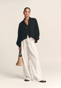 Črna srajca z dolgimi rokavi in gumbi, bele široke hlače, črne ravne sandale in tkan torbica. Gladek material in sproščen kroj.