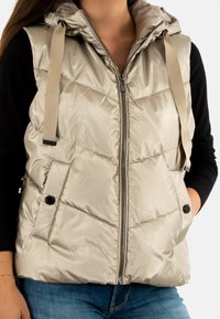 Veste matelassée beige clair avec capuche, fermeture éclair à l'avant, poches latérales et léger éclat. Présente des boutons noirs et des sangles en tissu.