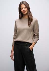 Ljusbeige tröja med rund halsringning och ribbade ärmslut, ihop med mörka rutig byxor. Tyget verkar mjukt med minimal textur.