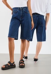 Denimshorts, mörkblå, knälånga, avslappnad passform, fem-fickors design, synliga sömdetaljer, kombinerade med svarta sandaler och skor.
