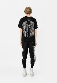 Camiseta negra con gráficos de alas plateadas en la parte posterior, combinada con joggers negros desgastados que presentan etiquetas laterales y acentos blancos, y usados con botas blancas.