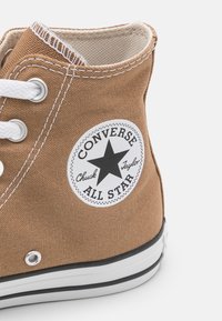 Brązowe wysokie trampki Converse All Star z bia łymi sznurówkami, widocznym szwem i czarną łatką z logo gwiazdy z boku.