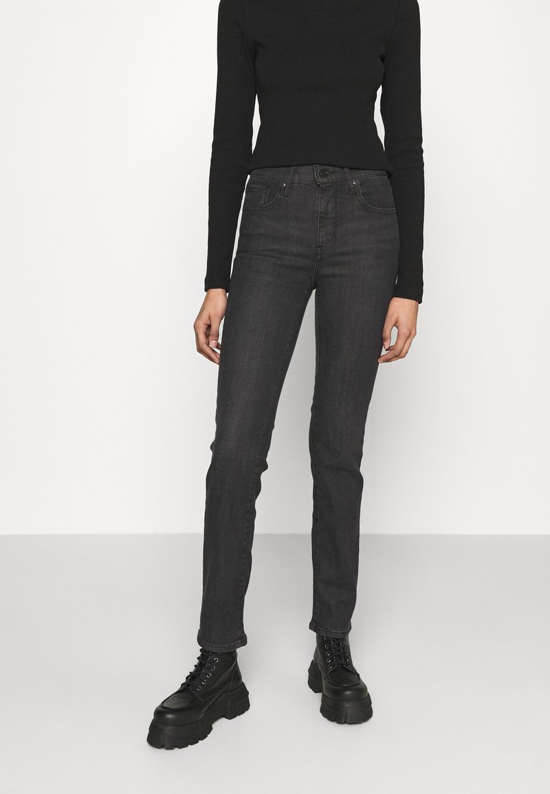 Personne portant un haut noir à manches longues, un jean taille haute gris foncé et des bottines noires épaisses à lacets, debout devant un fond uni.