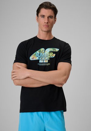 Młody mężczyzna z założonymi rękami, ubrany w czarny T-shirt z napisem "Summer of Code" i niebieskie szorty na jednolitym szarym tle.